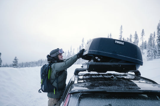 Person securing a Thule roof box on a car in a snowy landscape