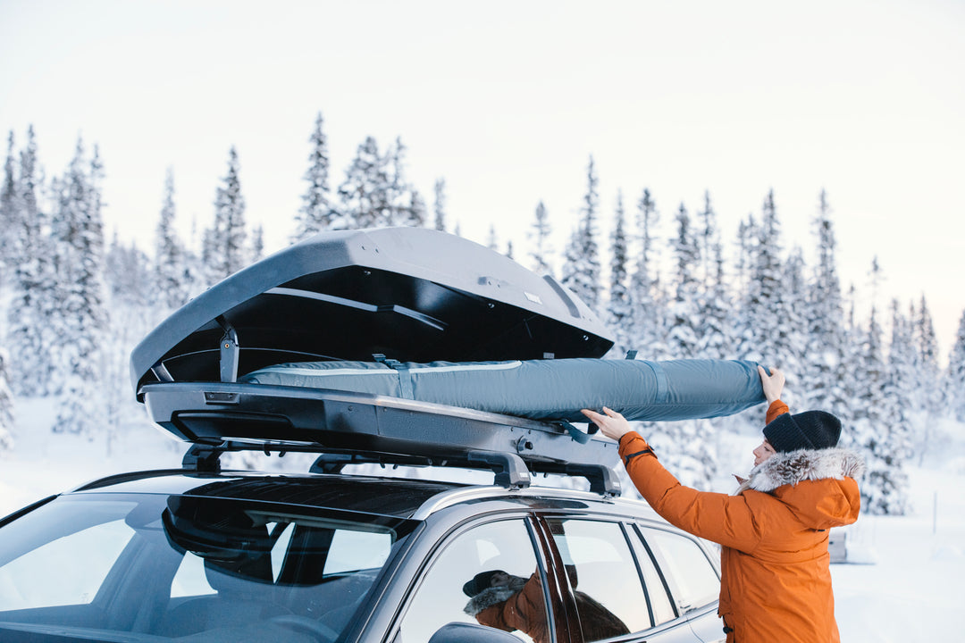 Person loading a snowboard onto a car roof rack in a snowy landscape.